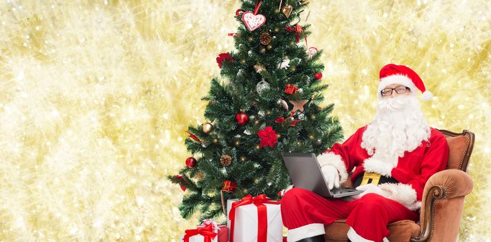 technology, holidays and people concept - man in costume of santa claus with laptop computer, gifts and christmas tree sitting in armchair over yellow lights background