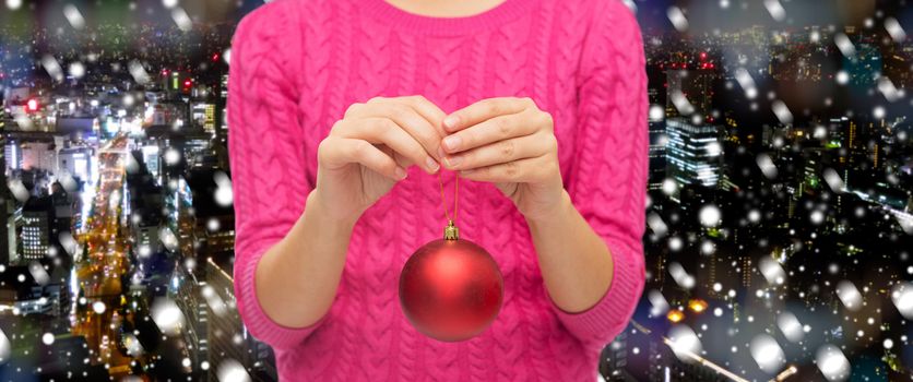 christmas, decoration, holidays and people concept - close up of woman in pink sweater holding christmas ball over snowy night city background
