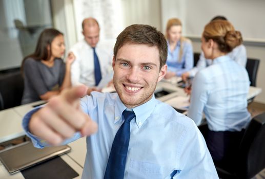 business, people, gesture and teamwork concept - smiling businessman pointing finger to you with group of businesspeople meeting in office