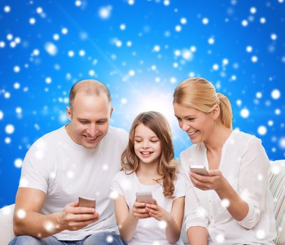 family, christmas holidays, technology and people - smiling mother, father and little girl with smartphones over blue snowy background