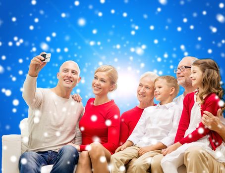 family, holidays, generation, christmas and people concept - smiling family with camera taking selfie and sitting on couch over blue snowy background