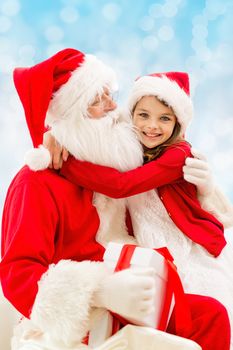 holidays, christmas, childhood and people concept - smiling little girl hugging with santa claus over blue lights background