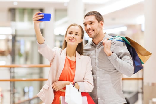 sale, consumerism, technology and people concept - happy young couple with shopping bags and smartphone taking selfie in mall