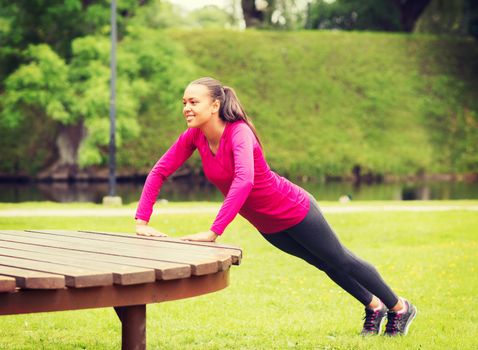 fitness, sport, training, park and lifestyle concept - smiling african american woman doing push-ups on bench outdoors