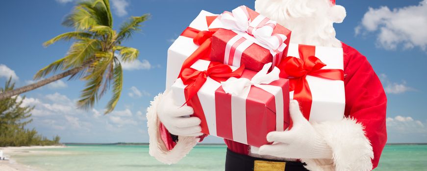 christmas, holidays, travel and people concept - close up of santa claus with gift boxes over tropical beach background