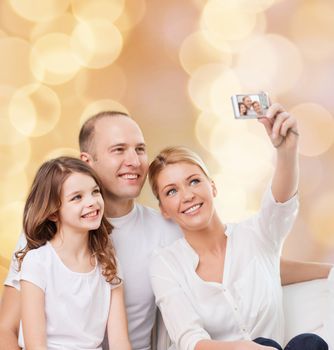 family, holidays, technology and people - smiling mother, father and little girl making selfie with camera over beige lights background
