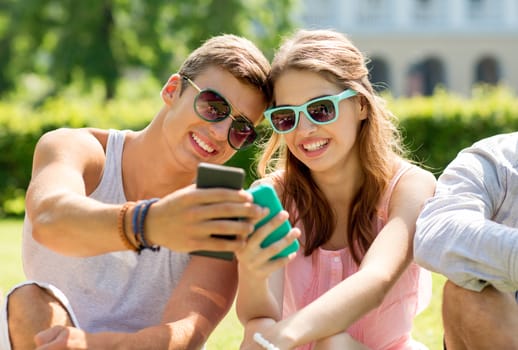 friendship, leisure, summer, technology and people concept - group of smiling friends with smartphone sitting on grass in park