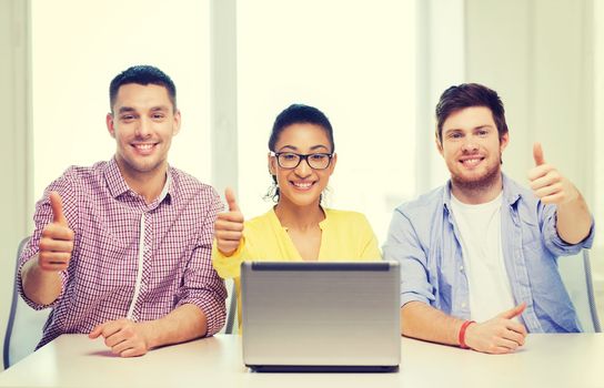 education, technology, business, startup and office concept - three smiling colleagues with laptop in office showing thumbs up