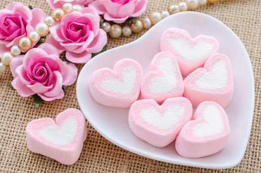Pink heart marshmallows in white bowl with rose flower on sack background.