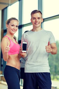 fitness, sport, advertising, technology and diet concept - smiling young woman and personal trainer with smartphone in gym showing thumbs up