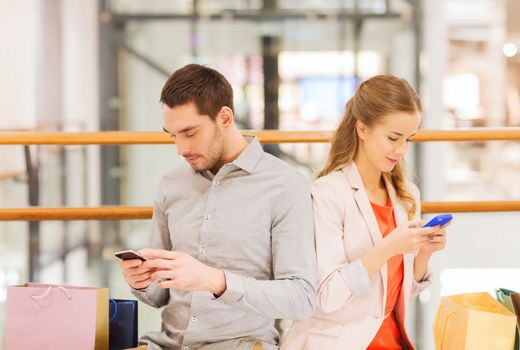 sale, consumerism, technology and people concept - young couple with shopping bags and smartphones in mall