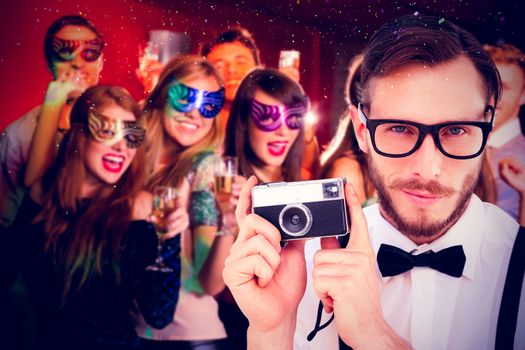 Geeky hipster holding a retro camera against friends in masquerade masks drinking champagne