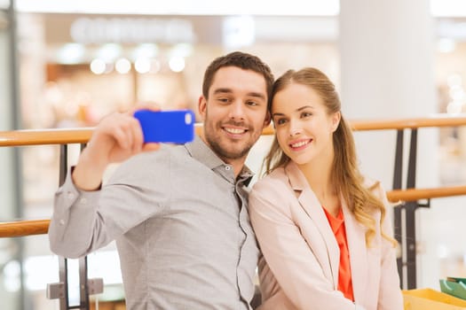 sale, consumerism, technology and people concept - happy young couple with shopping bags and smartphone taking selfie in mall