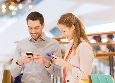 sale, consumerism, technology and people concept - happy young couple with shopping bags and smartphones in mall