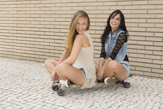 Two beautiful and young girlfriends having fun with a skateboard, in front of a brick wall