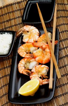 Delicious Asian Style Roasted Shrimps with Lemon and Chopsticks, Soy Sauce and Boiled Rice in Square Bowls  on Straw Mat background closeup