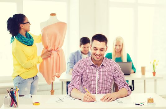 startup, education, fashion and office concept - smiling male drawing sketches and female adjusting dress on mannequin in office