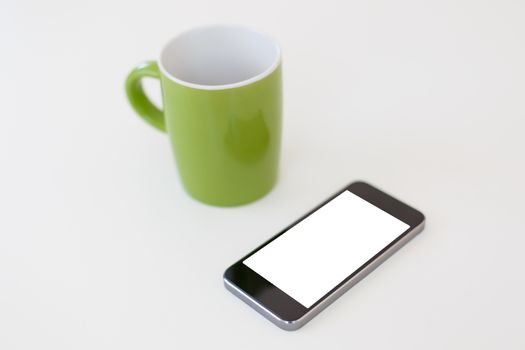 Close up detailed view of a smart phone, with white blank copy space screen, near a green mug, on white background.