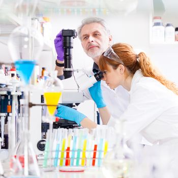 Attractive young female scientist and her senior male supervisor looking at the microscope slide in the life science research laboratory. Bichemistry, genetics, forensics, microbiology...