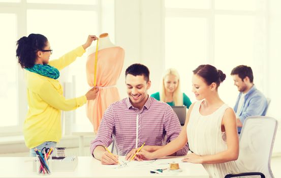 startup, education, fashion and office concept - smiling designers drawing sketches and adjusting dress on mannequin in office