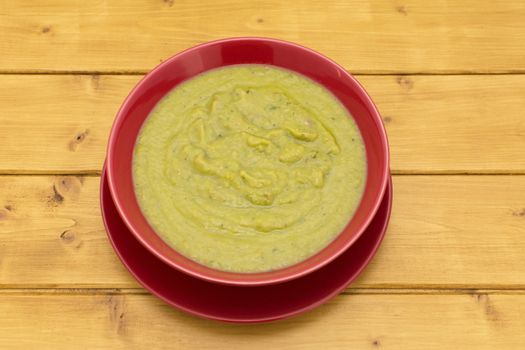 Bowl of green pea and ham soup in a red bowl on a wooden kitchen table