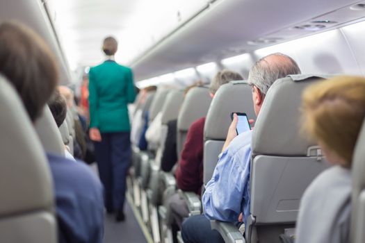 Interior of airplane with passengers on seats waiting to taik off. Stewardess in green uniform walking the aisle. Horizontal composition.