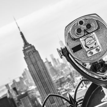 New York City, USA. Vintage tourist binoculars at Top of the Rock observation deck in front of Manhattan downtown skyline with Empire State Building and skyscrapers at sunset. Black and white.