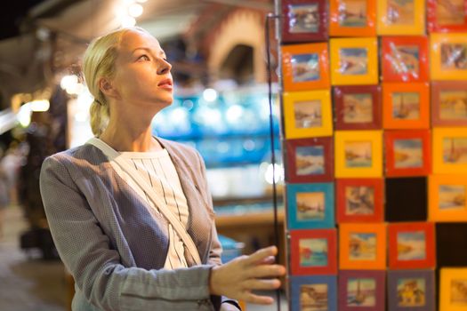 Beautiful casually dressed caucasian blond woman shopping for vacation souvenirs on crafts market in Chania, Crete, Greece.