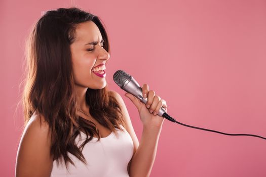 Portrait of a charming and beautiful woman singing with microphone in studio over pink background.