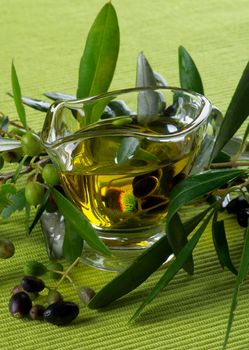 Arrangement of Olive Oil in Glass Gravy with Raw Green and Black Olives with Leafs closeup on Green Textile background