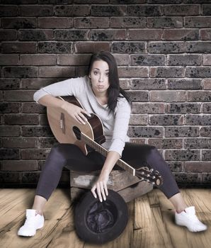 Young female busker sitting on a wooden box playing her guitar for money.