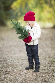 Baby Girl In Red Mittens and Cap Holding Small Christmas Tree Outdoors.