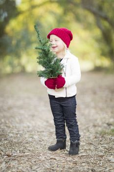 Baby Girl In Red Mittens and Cap Holding Small Christmas Tree Outdoors.