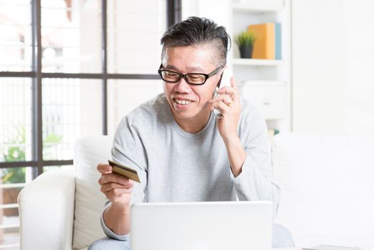Smiling Asian mature man making order by phone, looking at credit card, and using laptop computer at home.