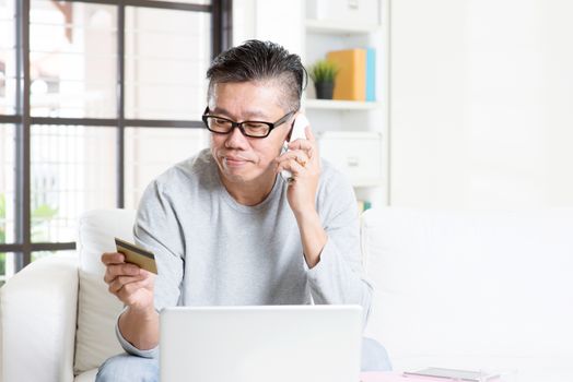 Asian mature man making order by phone, looking at credit card, and using laptop computer at home.