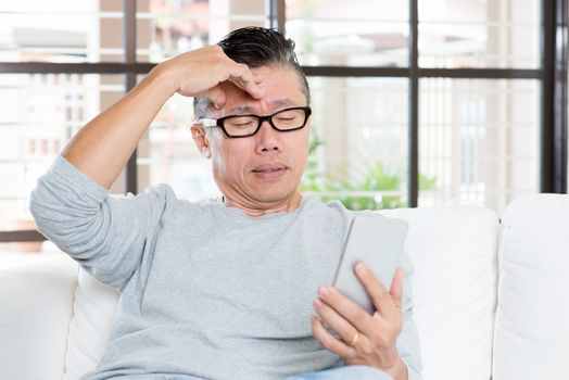 Portrait of 50s mature Asian man headache while using smart phone, sitting on sofa at home.