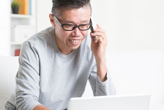 Portrait of 50s mature Asian man calling on phone while using computer internet working from home, sitting on sofa at indoors.