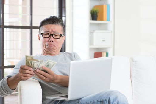 Financial problem concept. Portrait of 50s mature Asian man counting money with worried expression, sitting on sofa at home.