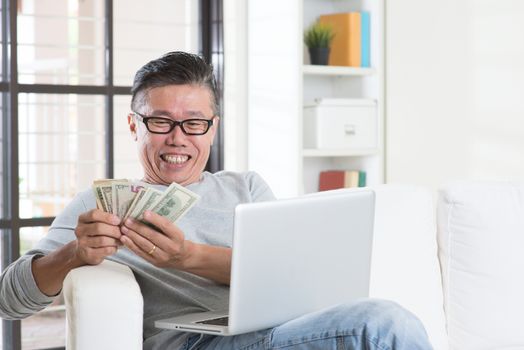 Portrait of happy 50s mature Asian man using internet computer and counting cash at home, earning money from his successful online business. Working from home concept.