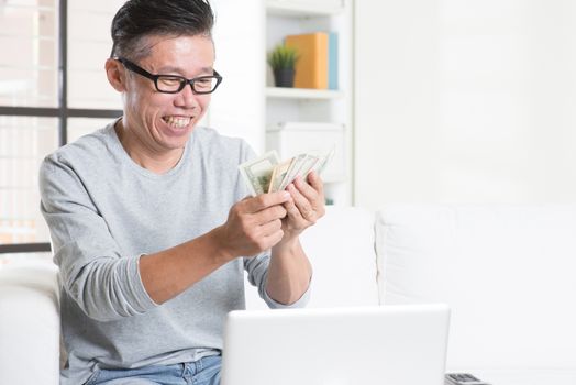 Portrait of 50s mature Asian man counting cash, earning money from his successful online business. Working from home concept.