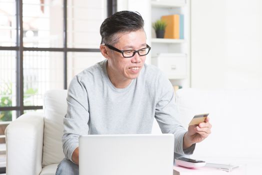 Portrait of 50s mature Asian man using computer doing online shopping with credit card, sitting on sofa at home, senior retiree indoors living lifestyle.