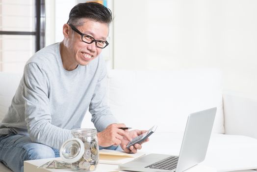 Mature 50s Asian man counting on money using calculator and laptop computer. Saving, retirement, retirees financial planning concept. 
