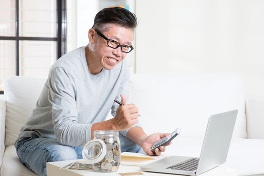 Mature 50s Asian man giving thumb up while counting on money using calculator and laptop computer. Saving, retirement, retirees financial planning concept. 