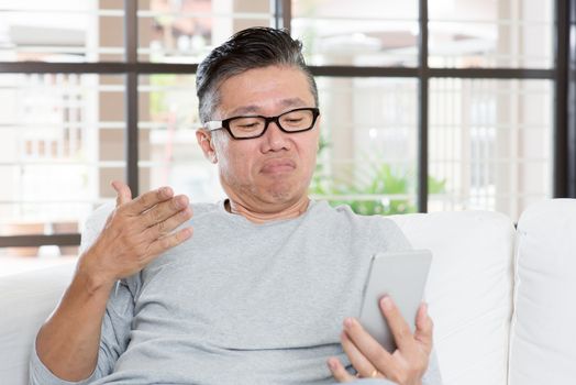 Portrait of 50s mature Asian man with negative face expression while using smart phone, sitting on sofa at home.