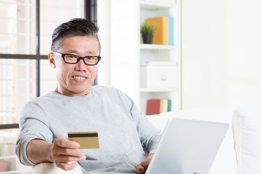 Portrait of 50s mature Asian man using computer paying bill with credit card, sitting on sofa at home, senior retiree indoors living lifestyle.