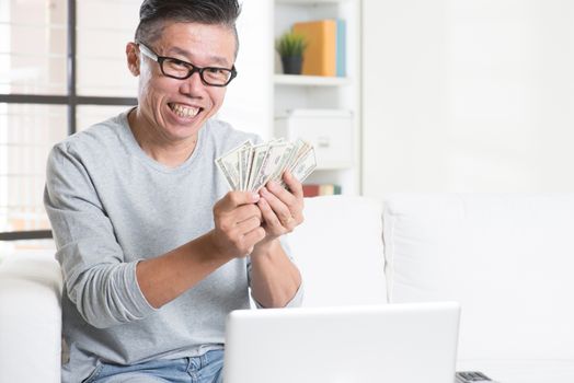 Portrait of 50s mature Asian man using internet computer and counting cash at home, earning money from his successful online business. Working from home concept.