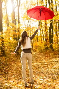 Beautiful cheerful young woman with red umbrella having fun in sunny forest in autumn colors.