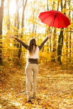 Beautiful cheerful young woman with red umbrella having fun in sunny forest in autumn colors.
