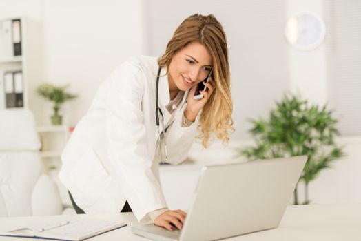 Beautiful young female doctor in her consulting room, using phone and writing on laptop.