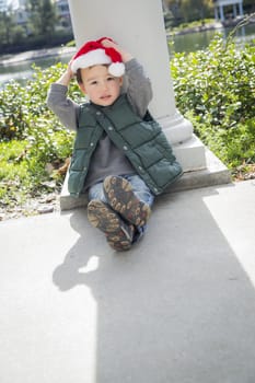 Cute Mixed Race Boy Sitting At The Park Wearing Christmas Santa Hat.